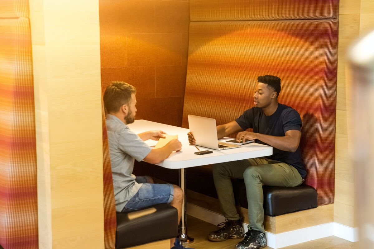Kaksi nuorta miestä istuu loosissa keskustelemassa, toisella tietokone.