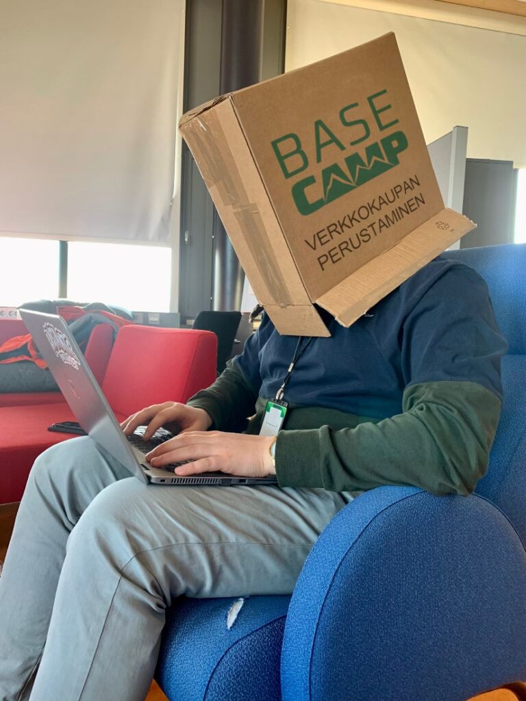 Henkilö istuu nojatuolissa kannettava tietokone sylissä ja päässään pahvilaatikko, jossa lukee "Base Camp - Verkkokaupan perustaminen".