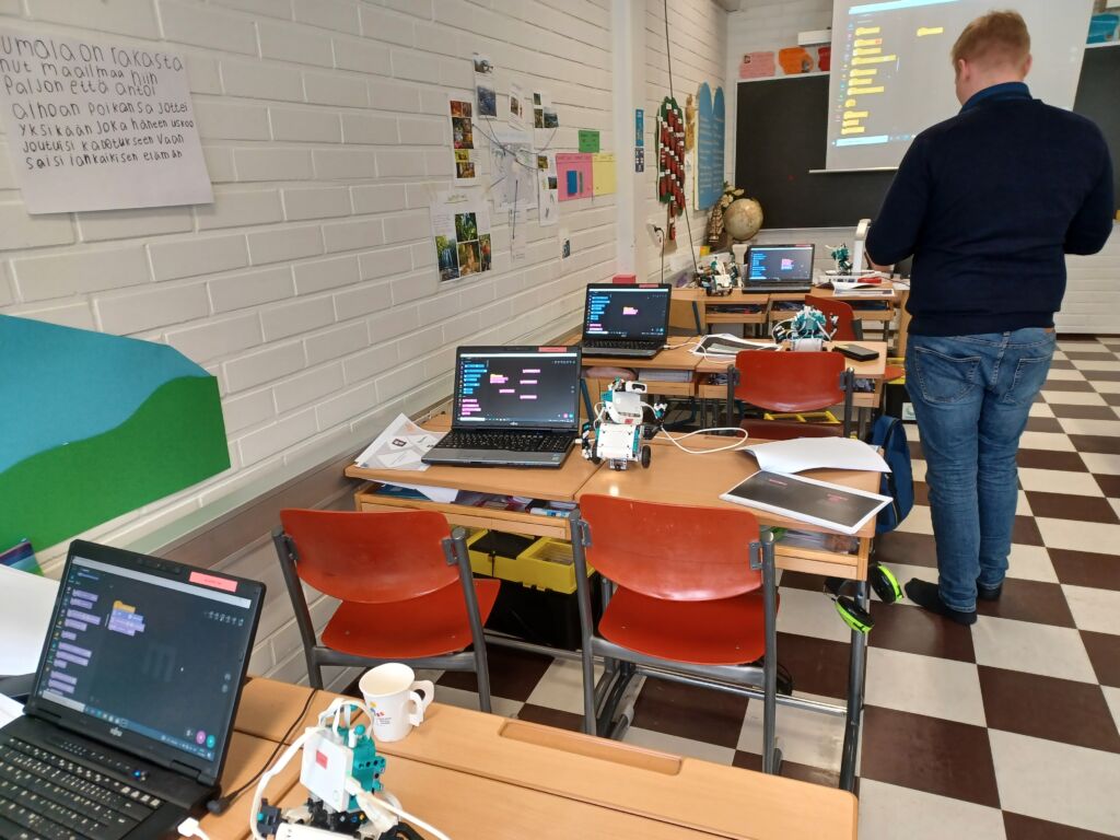Luokkatilassa pulpetteja tuoleineen, tietokoneita ja robotteja. Ohjaaja erään pulpetin vieressä selkä katsojaan päin.