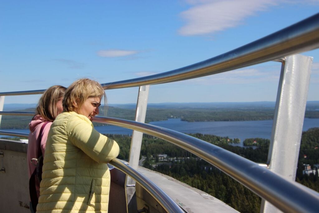 Kaksi naista seisoo korkealla näkötornissa, maisemassa järviä ja metsää.