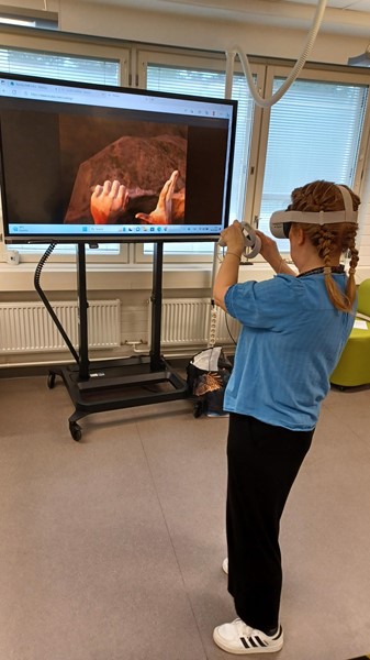 Nainen seisoo seinään kiinnitetyn näytön edessä. Naisella on päässä virtuaalilasit ja käsissä ohjaimet.