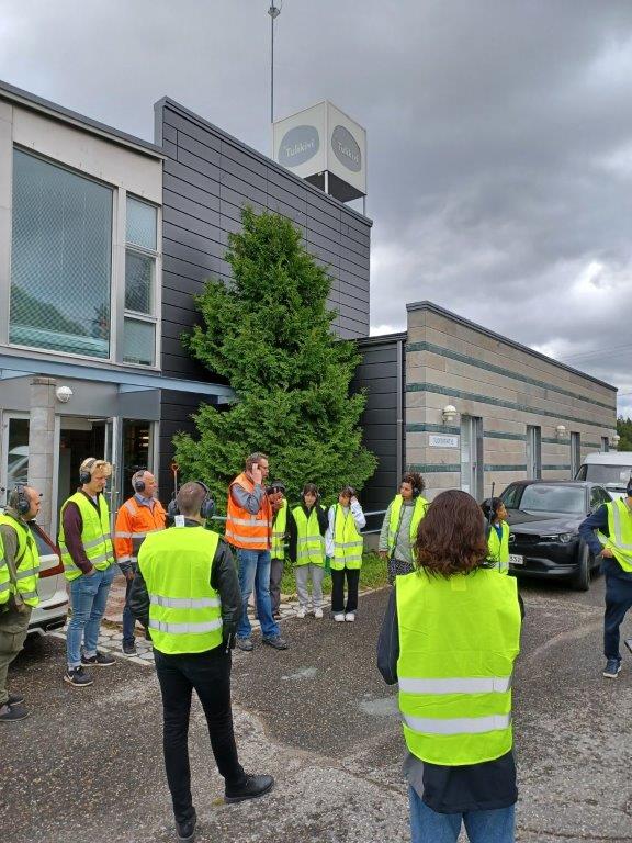 Tehdasrakennus jonka edustalla ihmisiä huomioliivit päällään