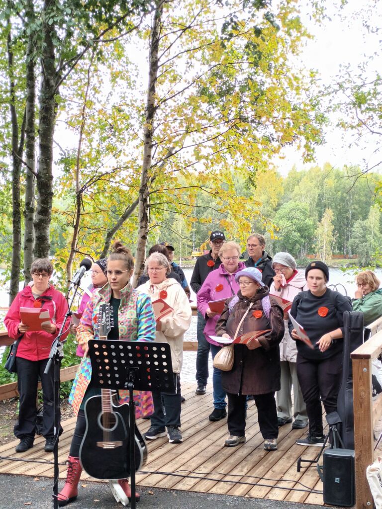 Muistista Laulajat -kuoro esiintymässä Kaivannonpuistossa. Taustalla koivuja ja Pielisjoki