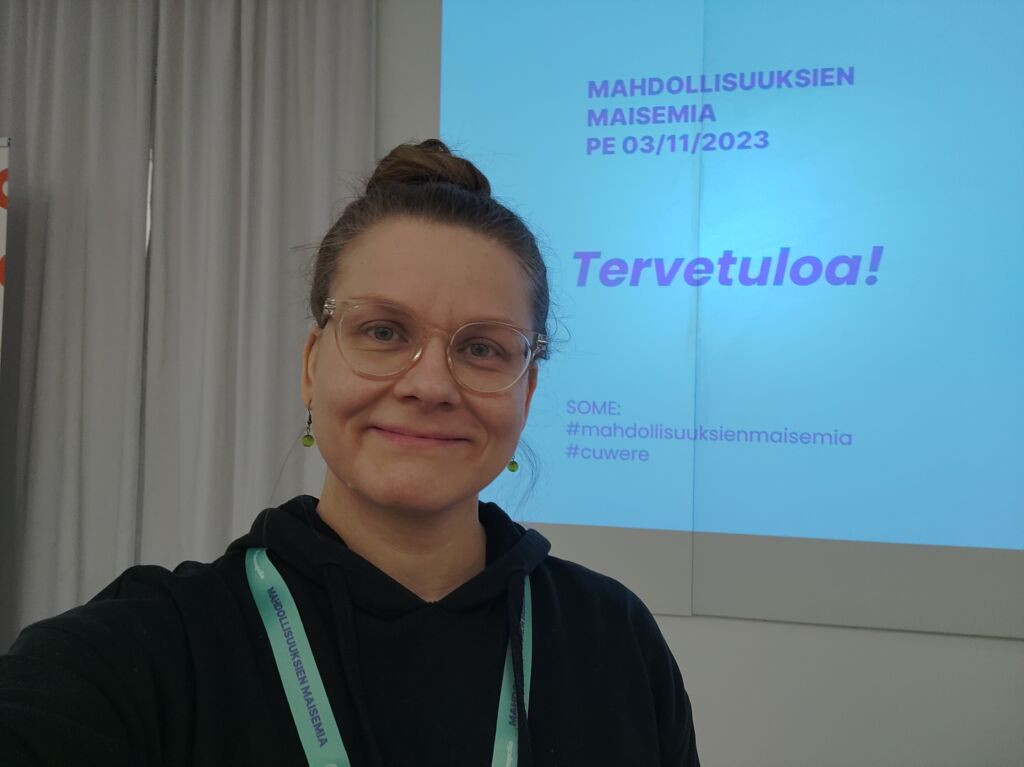 A slide projected on the wall welcoming to “Mahdollisuuksien maisemia” event. Maria Korkatti in front of the slide smiling to the camera.