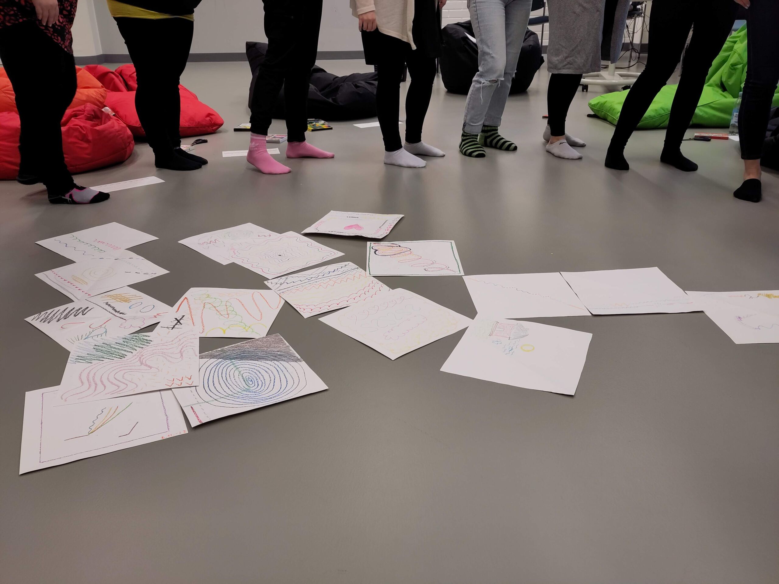 There are square-shaped papers with abstract line images on the floor. People are standing around them, only their legs can be seen.