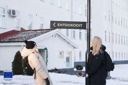 Kaksi naista ulkona valkoisen rakennuksen edessä talvivaatteissaan. Naisten keskellä viitta jossa lukee Ehtookodit.