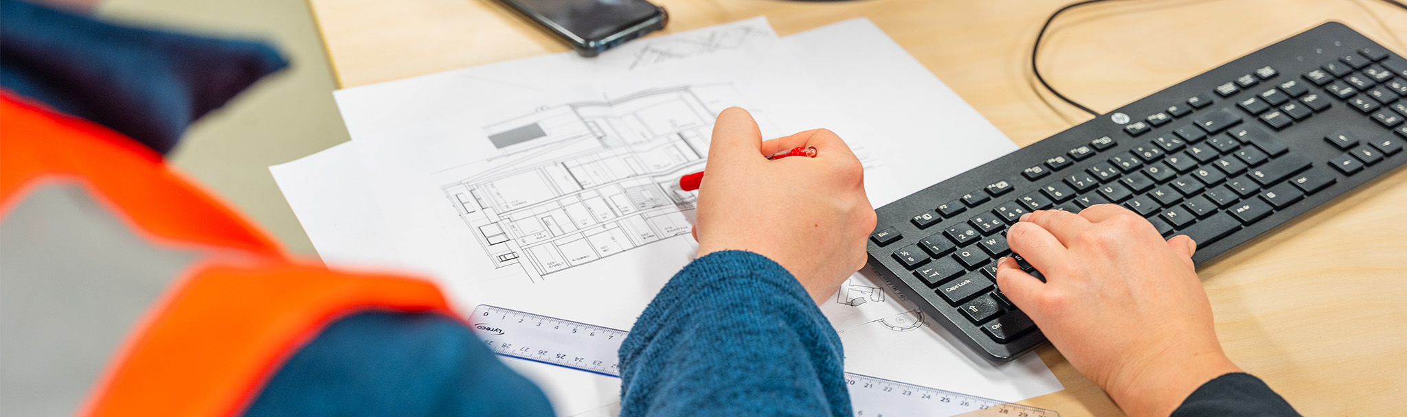 Henkilö osoittaa pöydällä olevaa rakennuspiirrustusta punaisella kynällä. Samalla toisen henkilön kädet kirjoittavat tietokoneen näppäimistöllä.