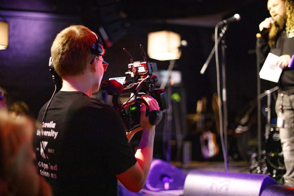 Poika kuvaa kameralla henkillöä joka seisoo lavalla mikrofoni kädessä