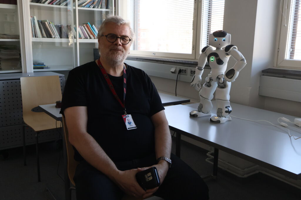 Mies istuu pöydän ääressä, pöydällä pienikokoinen robotti katsoo kameraan