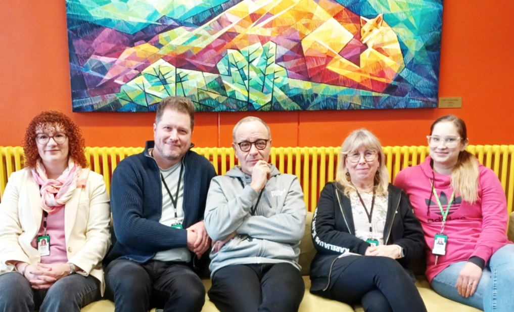 Viisi henkilöä istuu sohvalla vierekkäin, takana värikäs maalaus