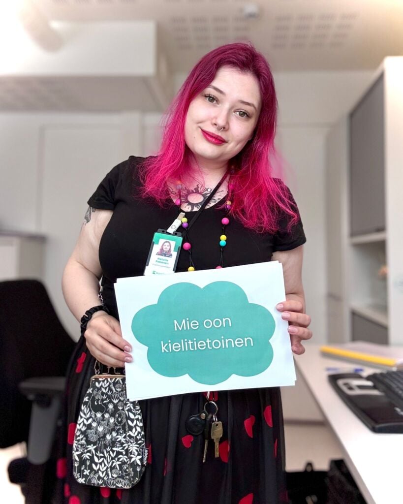 Punatukkainen nainen pitää kylttiä jossa lukee "Mie olen kielitietoinen"