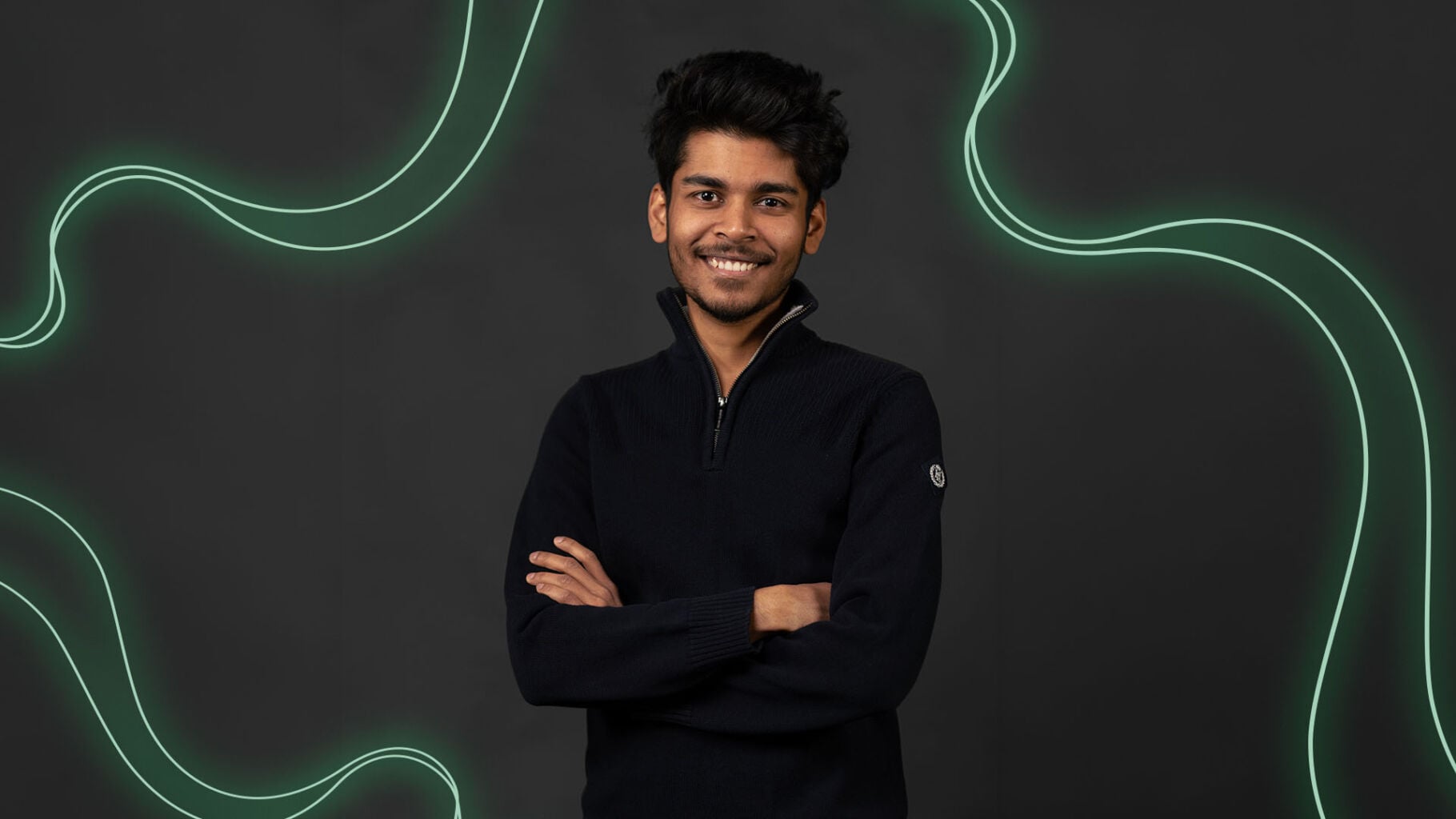 Portrait of a person standing with arms crossed, wearing a dark sweater. The background is a dark, charcoal-grey surface with green, wavy light lines running across it. The person is looking directly at the camera with the corners of the mouth slightly raised.