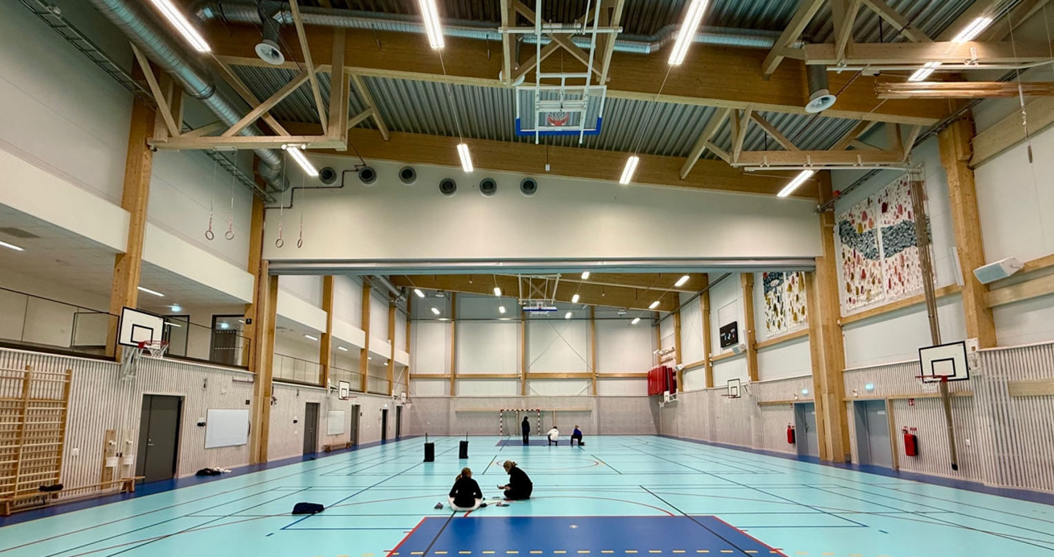 Urheiluhallin sisätilat. Sisäpelikentällä istuu muutamia ihmisiä. Kentän laidoilla on koripallokoreja. Katossa on puiset palkit.