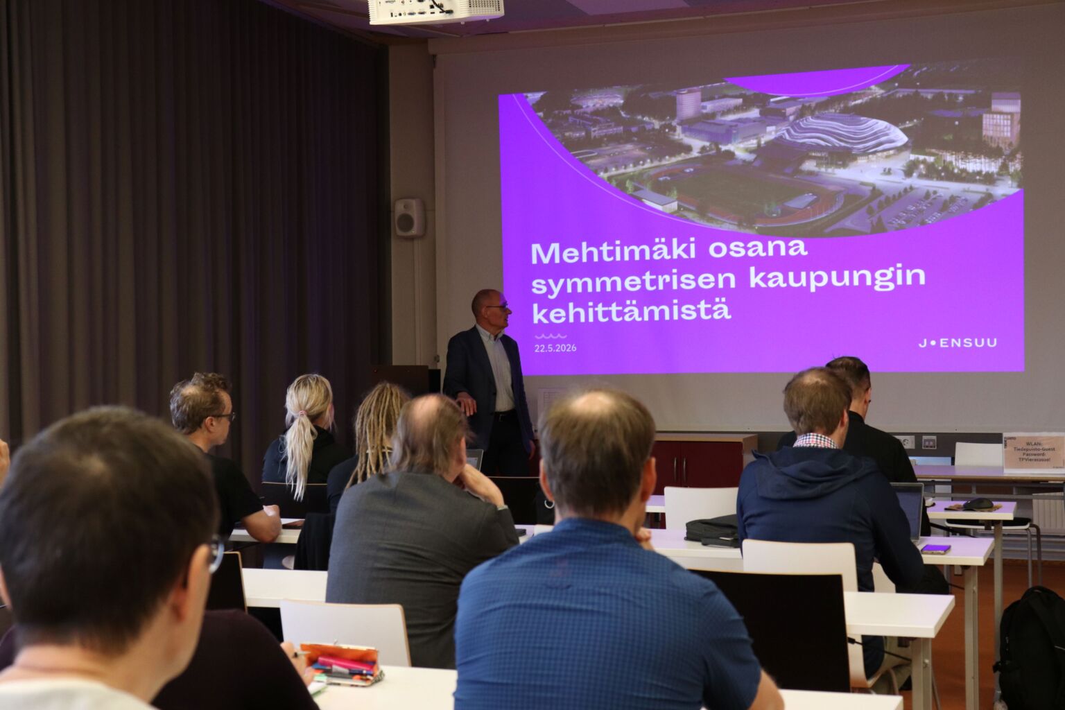 Mies luentosalin etuosassa, taustalla esityksen otsikko Mehtimäki osana symmetrisen kaupungin kehittämistä, Yleisöä kuuntelemassa pöytien ääressä.